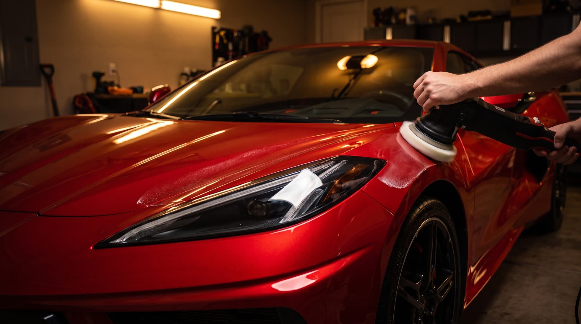 Dual-action polisher correcting the paint on the red sports car's front fender in a warm garage